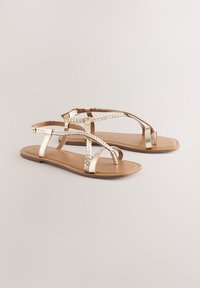 Sandali flat metallici dorati con cinturini intrecciati, suola liscia color marrone chiaro, design sottile e fibbia regolabile alla caviglia. Calzature estive casual.