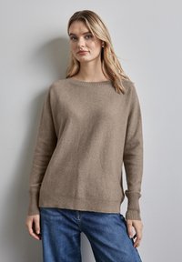 Beige gestrickter Pullover mit Rundhalsausschnitt, gerippten Bündchen und lässiger Passform. Getragen über blauen Jeans vor einem hellgrauen Hintergrund.