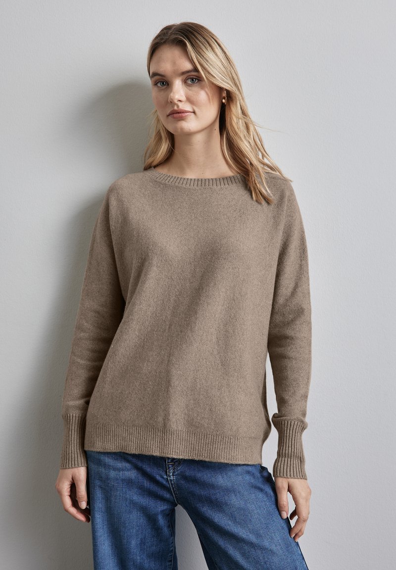 Beige gestrickter Pullover mit Rundhalsausschnitt, gerippten Bündchen und lässiger Passform. Getragen über blauen Jeans vor einem hellgrauen Hintergrund.