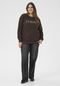 Usmievajúca sa žena v hnedom mikinovom svetri s nápisom "FEMME" a tmavosivými džínsami, stojí s jednou rukou držiacou lem mikiny.