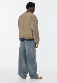 Giubbotto bomber beige con polsini e orlo a coste, abbinato a jeans in denim azzurro chiaro oversize con ampie tasche frontali e una vestibilità rilassata.