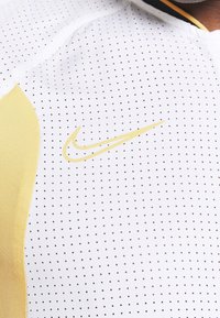 Gros plan sur un maillot de sport blanc à petits points noirs, avec des panneaux latéraux jaunes et un logo Nike swoosh jaune sur la poitrine.