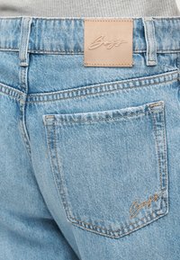 Ανοιχτό μπλε τζιν παντελόνι με πίσω τσέπη και καφέ δερμάτινη ετικέτα μάρκας με την επιγραφή "Boss", φορεμένο με ανοιχτό γκρι μπλουζάκι.