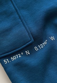 Donkerblauwe stof met een gestructureerde breiwerk en witte tekst: "51.5072° N 0.1276° W" gepositioneerd op een pocketdetail.
