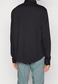 Schwarzes Langarmhemd mit Kragen, glatter Textur und geknöpften Manschetten, kombiniert mit hellblauen Hosen. Ansicht von hinten.