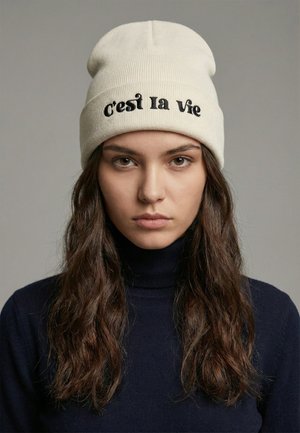 Giovane donna con lunghi capelli castani che indossa un berretto color crema ricamato con la scritta "c'est la vie" e un maglione a collo alto scuro.