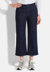 Pantalons en denim bleu foncé à jambes larges avec une fermeture à bouton et zip à l'avant, associés à des baskets avec des accents en argent. Texture lisse, design décontracté.