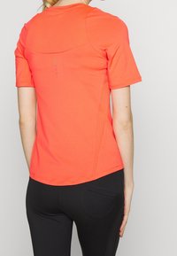 Nainen, jolla on päällään kirkkaan oranssi lyhythihainen urheiluvaate ja mustat tiukasti istuvat housut, näkyy selkäpuolelta yksinkertaista vaaleata taustaa vasten.