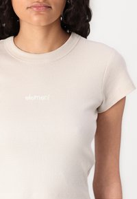 Beige ribbed t-shirt with short sleeves and a round neckline. Features a small, white, embroidered brand logo on the chest.