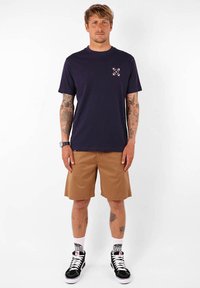 T-shirt à manches courtes navy avec un petit graphique multicolore sur la poitrine, associée à un short beige, des baskets montantes noires et des chaussettes à motif.