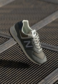 Sneaker New Balance grigio e verde oliva realizzata con materiali in rete e suede, caratterizzata da un grande logo "N" grigio e dettagli testurizzati.