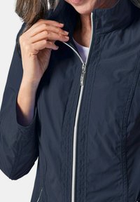 Marineblaue leichte Jacke mit hohem Kragen, silbernem Reißverschluss und dezenten Stickdetails. Verfügt über eine glatte Textur und lange Ärmel.