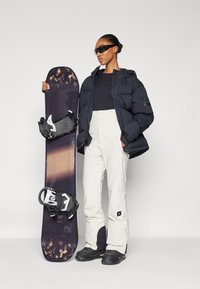 Black puffer jacket, black shirt, and white snow pants. Holding a dark snowboard with bindings. Sleek black sunglasses complete the look.
