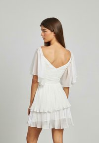Weißes Kleid mit geschichtetem, plissiertem Design, V-Ausschnitt auf der Rückseite und transparenten Schmetterlingsärmeln. Der Stoff scheint leicht und strukturiert zu sein.