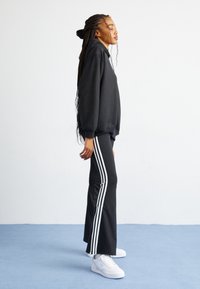 Zwarte hoodie met wijde mouwen, gecombineerd met zwarte broek met drie witte verticale strepen. Witte sneakers maken de outfit af.