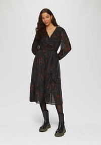 Schwarz gemustertes Wickelkleid mit langen Ärmeln, Volant-Saum und einem Bindegürtel; hat vertikale Streifen und eine transparente Textur. Kombiniert mit schwarzen Knöchelstiefeln.