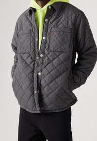 Prošívaná černobílá kostkovaná bunda se zapínáním na patentky, s dvěma náprsními kapsami a zaobleným lemem, vrstvená přes jasně neonovou mikinu s kapucí.