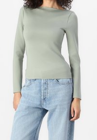 Donna che indossa un top a maniche lunghe verde chiaro aderente, infilato in un paio di jeans denim blu chiaro larghi, in piedi davanti a uno sfondo bianco.