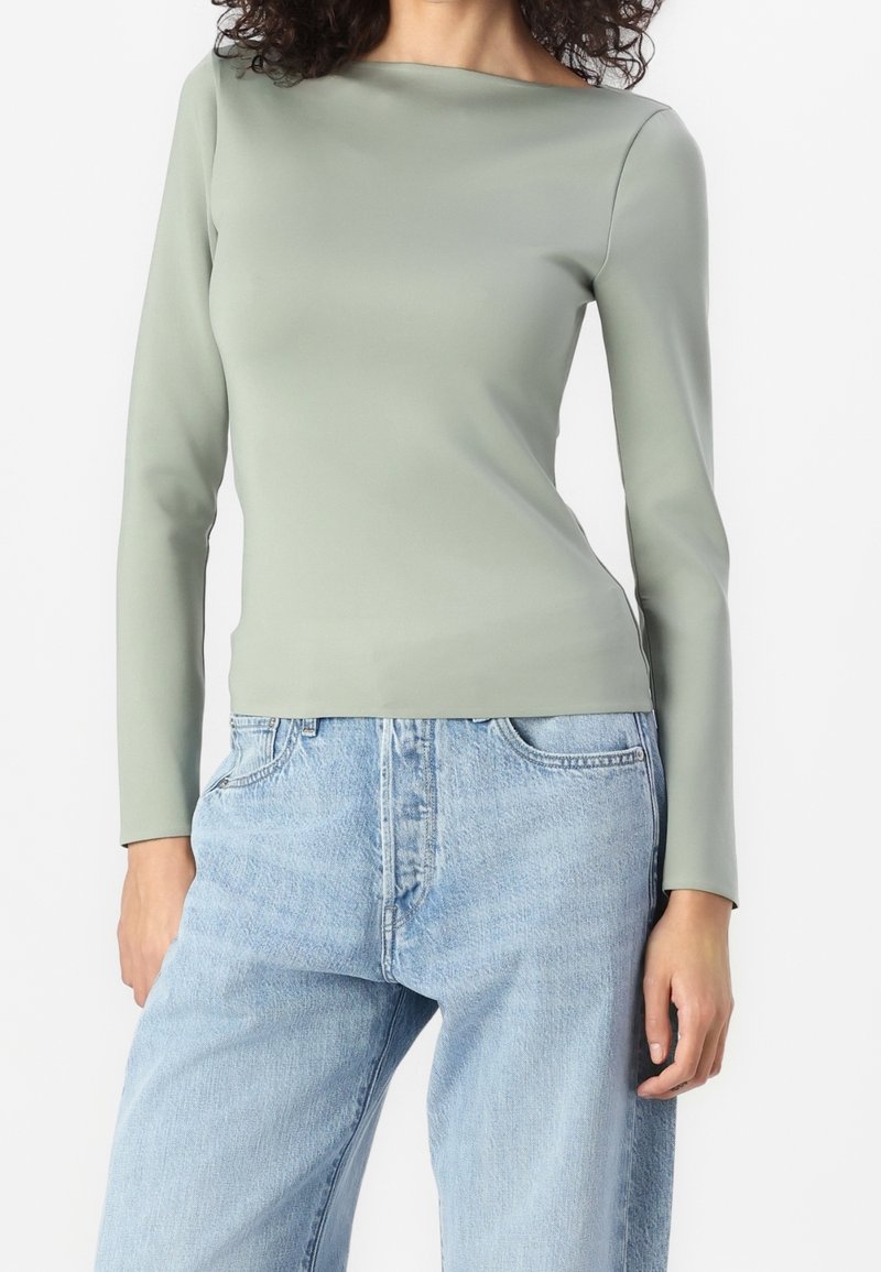 Donna che indossa un top a maniche lunghe verde chiaro aderente, infilato in un paio di jeans denim blu chiaro larghi, in piedi davanti a uno sfondo bianco.