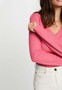 Femme dans un pull côtelé rose à manches longues croisant les bras sur la poitrine, portant un pantalon blanc taille haute avec un bouton doré et des clous.