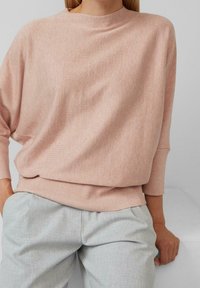Femme portant un pull à manches longues rose clair avec un ourlet côtelé et un pantalon plissé gris clair, assise avec une main posée sur une surface blanche.