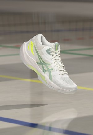 Bijela i svijetlo zelena Asics tenisica za trčanje vise iznad sportskog terena s obojenim linijama i sjenom ispod.