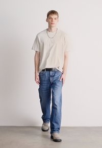 Ανοιχτό μπεζ πουκάμισο κοντομάνικο με ανάγλυφο λογότυπο, συνδυασμένο με μπλε τζιν. Το μοντέλο φοράει γκρι παπούτσια και μαύρη ζώνη, στέκεται μπροστά από ένα απλό φόντο.