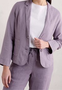 Blazer en lin violet clair avec une fermeture à un seul bouton, des revers crantés et deux poches, associé à un t-shirt blanc et un pantalon assorti.