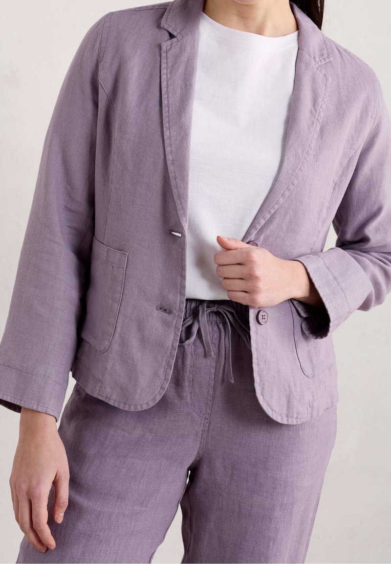 Blazer en lin violet clair avec une fermeture à un seul bouton, des revers crantés et deux poches, associé à un t-shirt blanc et un pantalon assorti.