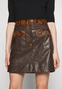 Brun läder A-linjeformad kjol med ormskinnsmönstrad midja, två framfickor med ormskinnsflikar och dekorativa knappar framtill.