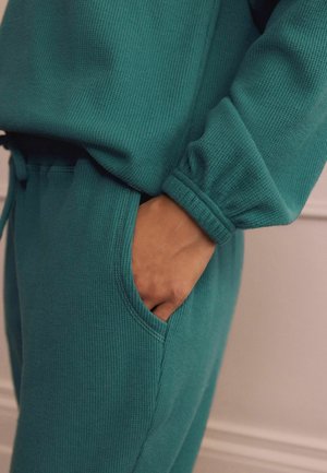 Teal textuur loungewear set met een losvallende top en broek met elastische boorden en zijzakken. De stof lijkt zacht en comfortabel te zijn.
