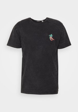 Mand iført sort t-shirt med lille grafik, off-white bukser og sort-blå sneakers, står på trægulv mod en hvid væg.