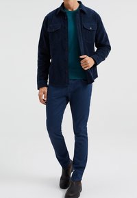 Navy corduroy shirt jacket with two chest pockets, paired with a teal sweater and dark blue pants. Black slip-on shoes complete the outfit.