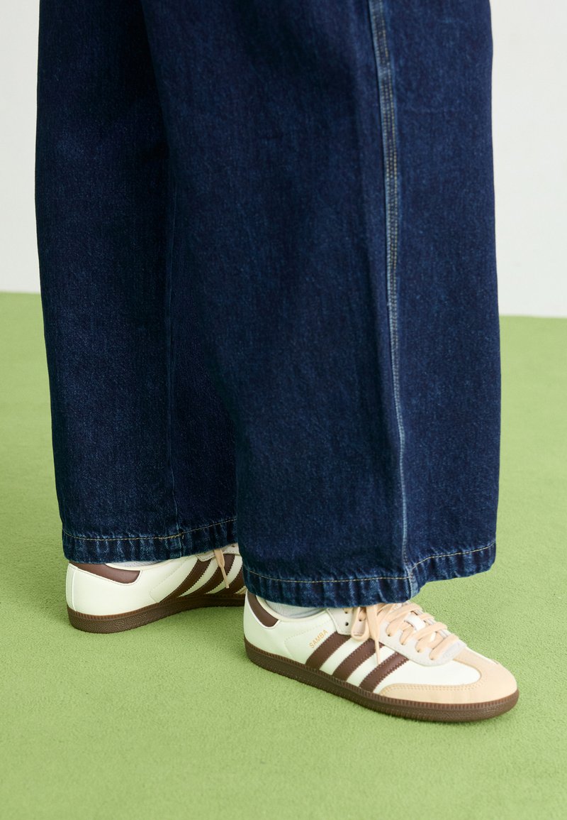 Sneakers bianchi e marroni con tre strisce, suola in gomma e lacci arancioni chiari; abbinati a pantaloni in denim blu scuro a vestibilità ampia.