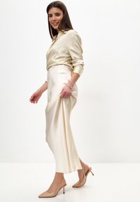 Blusa de seda beige con cuello y falda de satén beige a juego. La modelo lleva zapatos de tacón de punta con tiras traseras y adornos.
