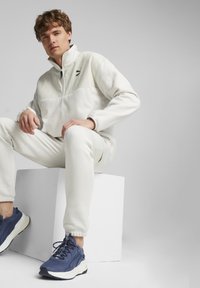 Giacca in pile bianca con chiusura a metà zip e logo nero, abbinata a jogger bianchi e sneakers blu scuro. Dettagli testurizzati visibili.