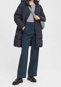 Abrigo negro acolchado con capucha, camiseta de rayas, pantalones azul turquesa y zapatos negros brillantes. Los accesorios incluyen un bolso cruzado verde.
