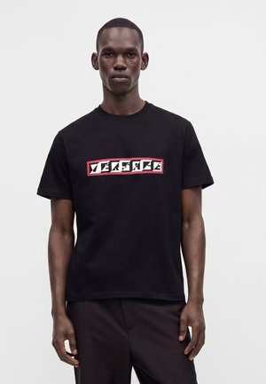 Homme portant un T-shirt noir Versace avec un logo blanc et rouge, debout devant un fond clair uni.