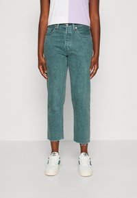 Persona con jeans teal cortos, zapatillas blancas con rayas verdes y una camiseta bicolor blanca y lavanda, de pie contra un fondo liso.