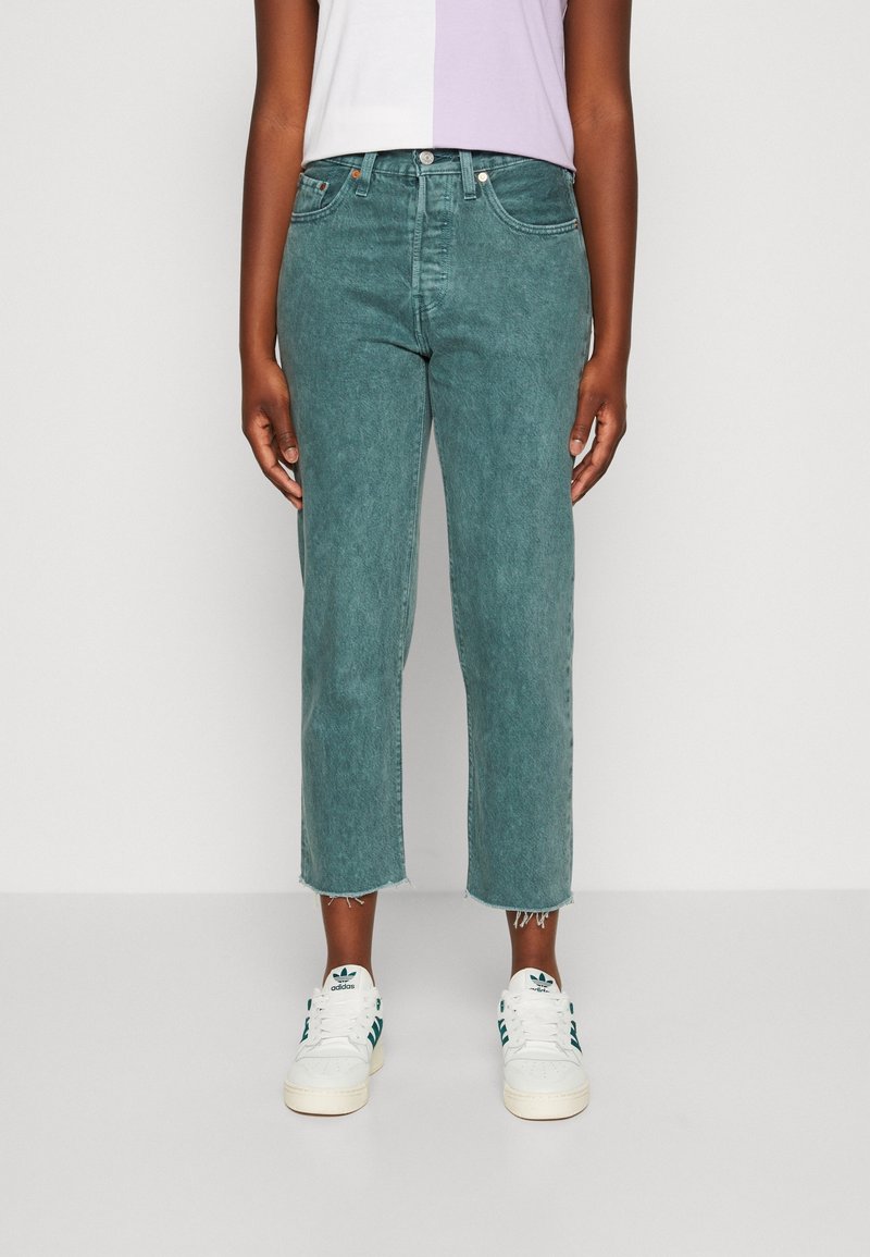 Persona con jeans teal cortos, zapatillas blancas con rayas verdes y una camiseta bicolor blanca y lavanda, de pie contra un fondo liso.