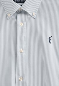 Ljusblå randig skjorta med knappar i bomull. Har vita knappar och en liten marinblå logotyp med en figur som håller en klubba på bröstet.