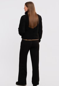 Giacca in pile nera con colletto alto, materiale testurizzato e orlo beige. Indossata con pantaloni larghi neri e calzature scure. Capelli lunghi visibili.