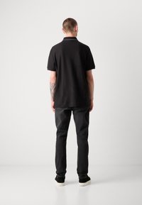 Svart kortärmad polo med enfärgad design, kombinerad med mörka jeans och svarta skor, sedd bakifrån.