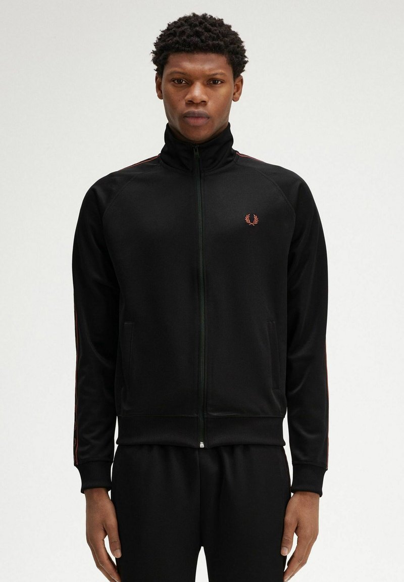 Giacca da track nera con zip e colletto alto, caratterizzata da strisce rosse sui maniche e un piccolo logo sul petto. Texture liscia.