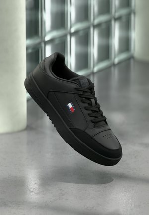 Czarny sneaker Tommy Hilfiger z logo po boku, czarnymi sznurówkami i perforowanym przodem, unoszący się wewnątrz na szarej polerowanej podłodze.