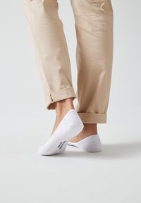 Chaussettes blanches à tige basse avec une texture côtelée, associées à un pantalon beige à revers, mettant en valeur la zone de la cheville et le logo de la chaussette au niveau du talon.