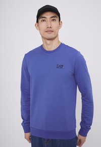 Felpa a maniche lunghe blu realizzata in un mix di cotone. Presenta un collo a giro e il logo EA7 nero sul lato sinistro del petto. Tessuto morbido, vestibilità rilassata.