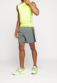 Neon-gelbes ärmelloses Sporttop, graue Shorts mit schwarzem Seitenstreifen und hellgrüne Laufschuhe mit orangefarbenen Akzenten.