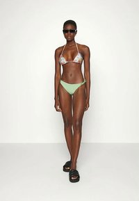 Mulher com top de biquíni halter floral, parte de baixo de biquíni verde claro com amarração lateral, sandálias slide pretas e óculos de sol pretos, de pé contra um fundo simples.