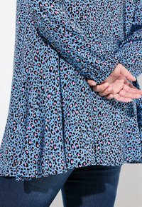 Dlouhý rukáv, modrý top s leopardím vzorem s černými a růžovými skvrnami, s plisovaným designem na lemu, spárovaný s tmavomodrými džíny.